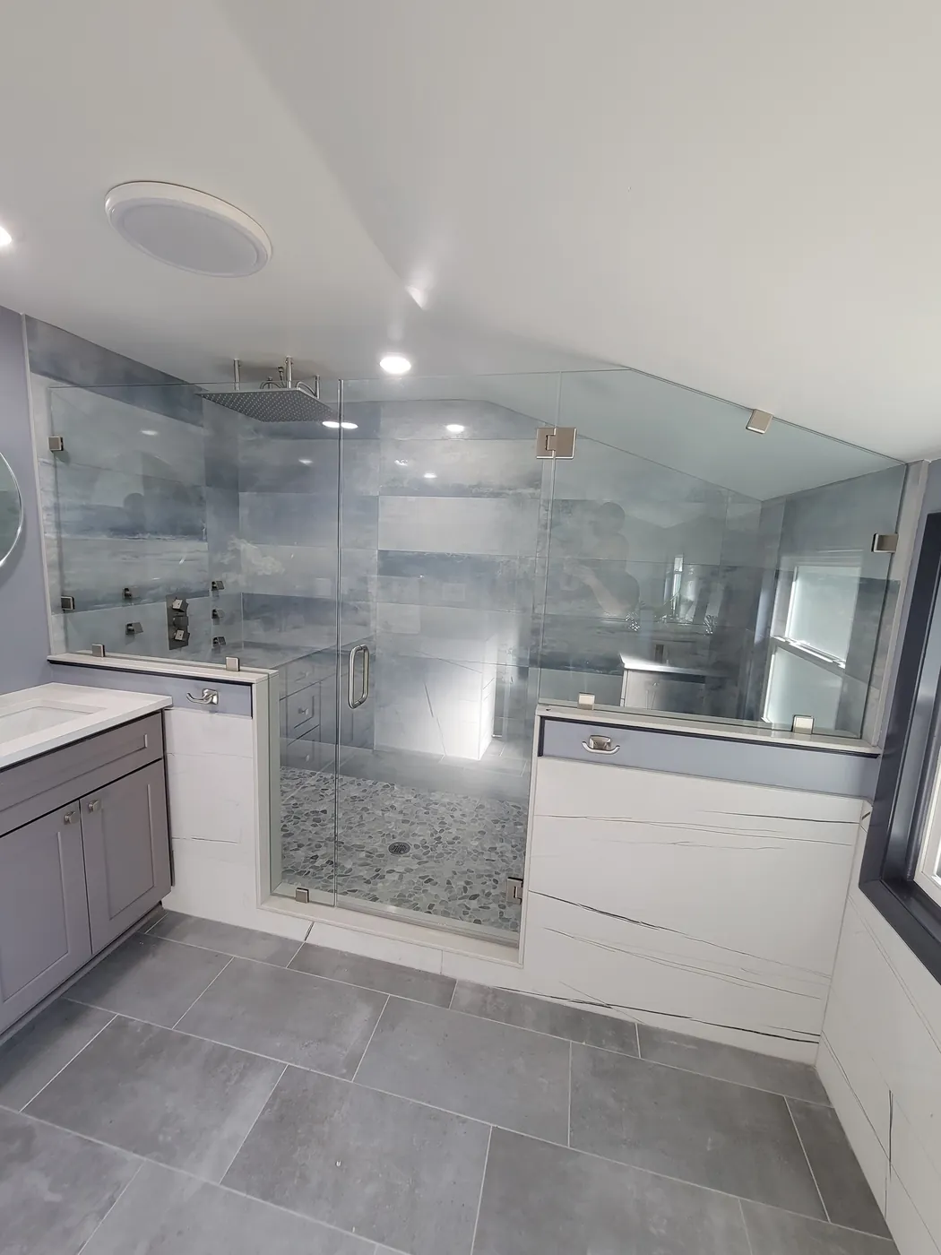 A large frameless glass shower enclosure over two tiled knee walls, with a pebble floor and blue-grey stone tile