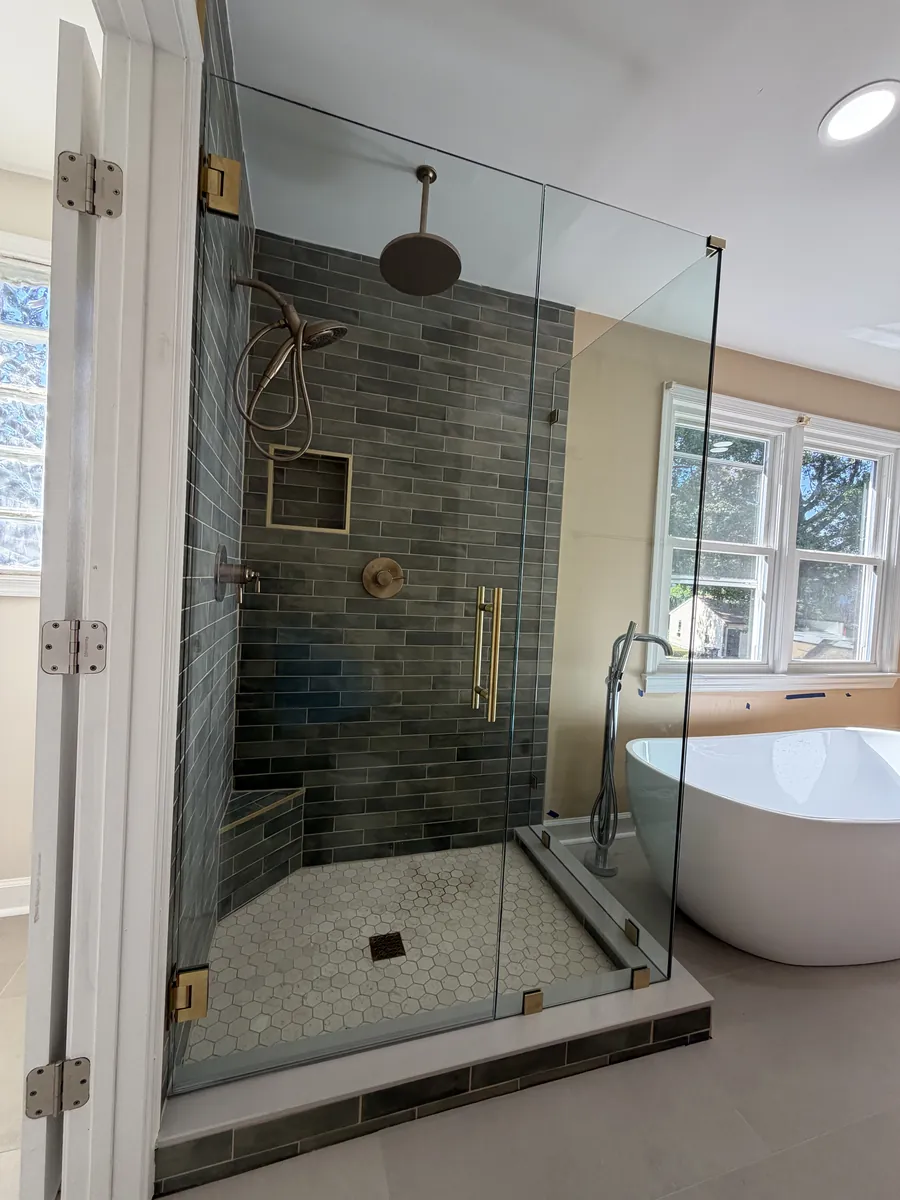 A corner frameless shower with gold hardware and green-grey tile, a freestanding tub beside it